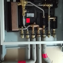 Hydro-Instal - Węzeł cieplny w obudowie z widocznymi rurami izolowanymi, oznaczonymi czerwoną taśmą i literami R i V, część instalacji grzewczej.