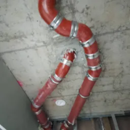 Czerwone rury instalacyjne z metalowymi obejmami, zamontowane pod surowym betonowym sufitem w trakcie prac wykończeniowych.