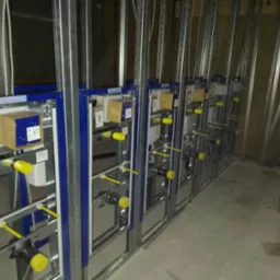 Szereg podtynkowych stelaży WC w trakcie montażu, z widocznymi metalowymi konstrukcjami, niebieskimi obudowami, żółtymi elementami i rurami, w surowym wnętrzu.