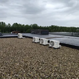 Cztery zewnętrzne jednostki klimatyzacyjne Panasonic zamontowane na płaskim dachu pokrytym żwirem, widok z góry w pochmurny dzień.