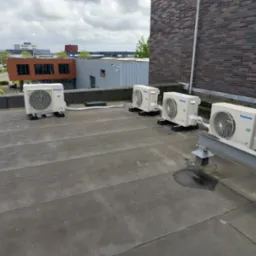 Cztery jednostki zewnętrzne klimatyzacji Panasonic zamontowane na płaskim dachu budynku z widokiem na zabudowania miejskie w pochmurny dzień.