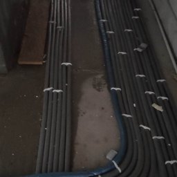 Hydro-Instal - Rzędy szarych i niebieskich rur, zabezpieczonych białymi opaskami, ułożone równolegle na betonowej podłodze w surowym wnętrzu. Ślady butów widoczne na podłodze.