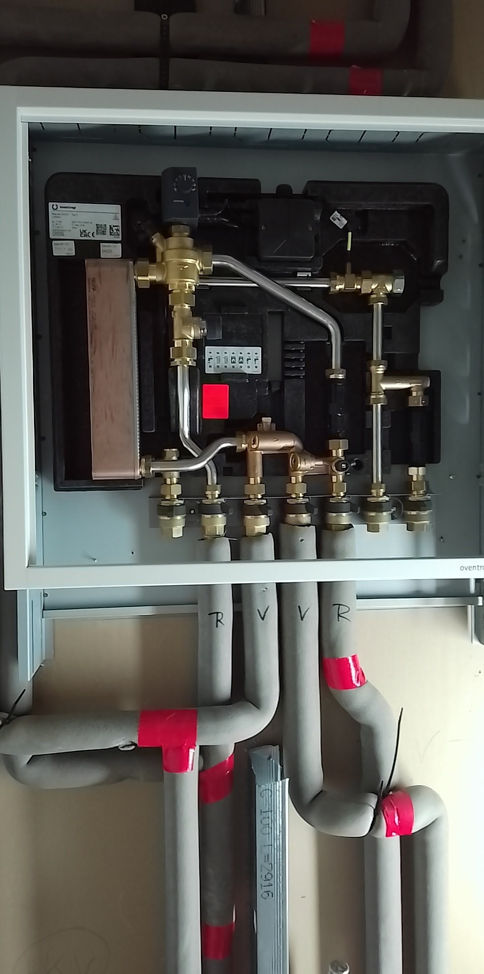 Węzeł cieplny w obudowie z widocznymi rurami izolowanymi, oznaczonymi czerwoną taśmą i literami R i V, część instalacji grzewczej.