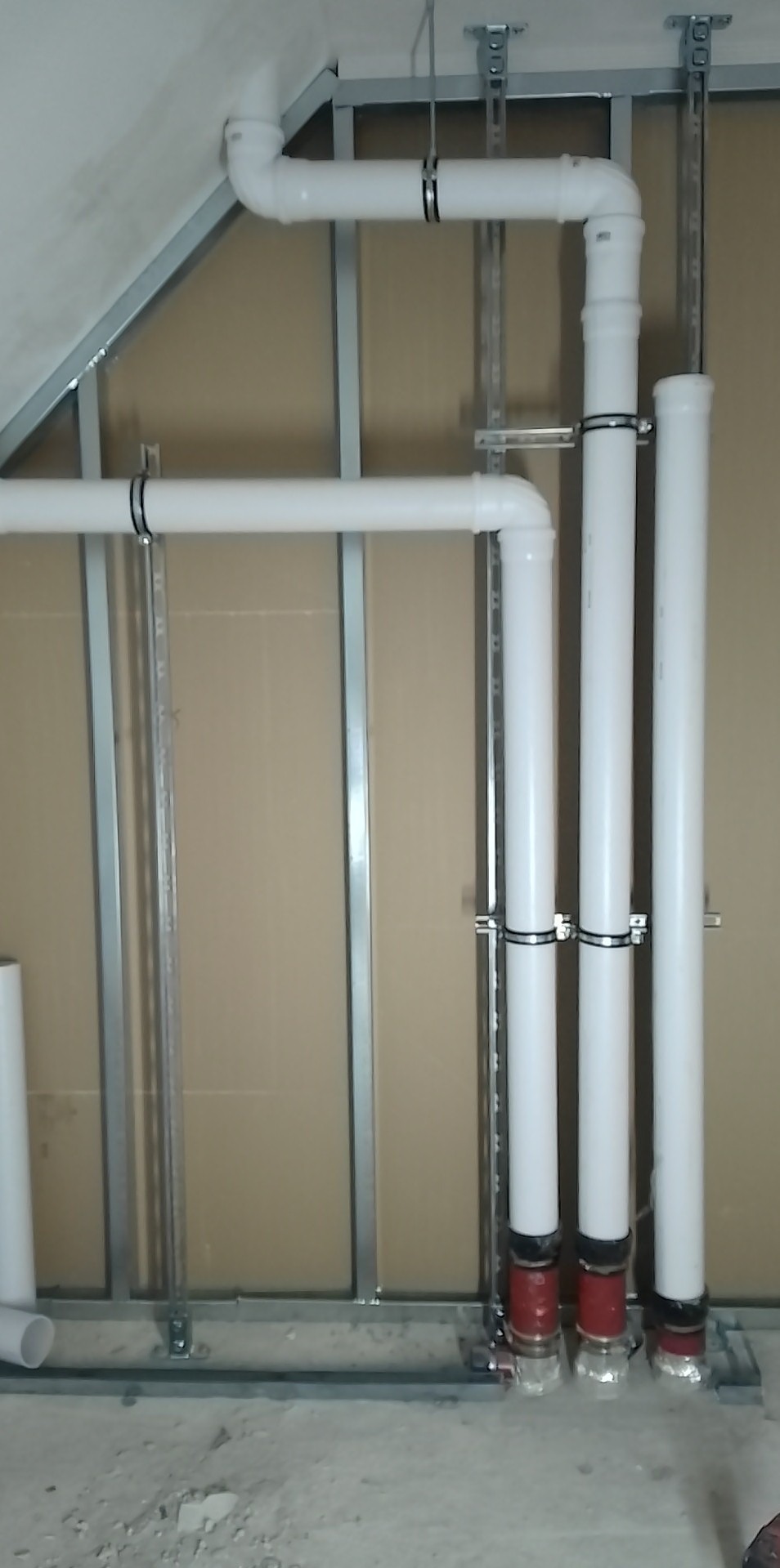 Białe rury kanalizacyjne zamocowane do ściany z płyt kartonowo-gipsowych na metalowym stelażu, widoczne obejmy mocujące i czerwone uszczelki u dołu rur.