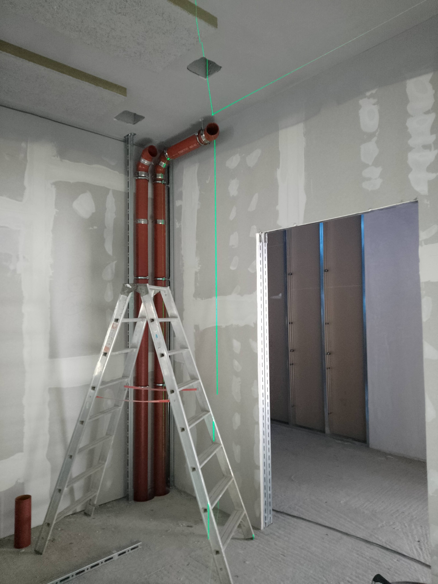 Wewnętrzne rury wentylacyjne z czerwonego tworzywa sztucznego, zamontowane przy ścianie na metalowej konstrukcji, widoczne z drabiny w surowym wnętrzu z płyt kartonowo-gipsowych i zielonym laserem...