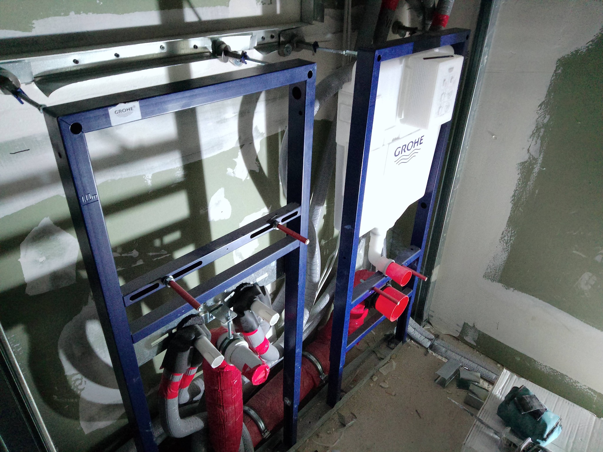 Dwa nieotynkowane stelaże podtynkowe WC marki Grohe, z widocznymi rurami doprowadzającymi wodę i odpływowymi, zamontowane przy surowej ścianie w trakcie prac instalacyjnych.