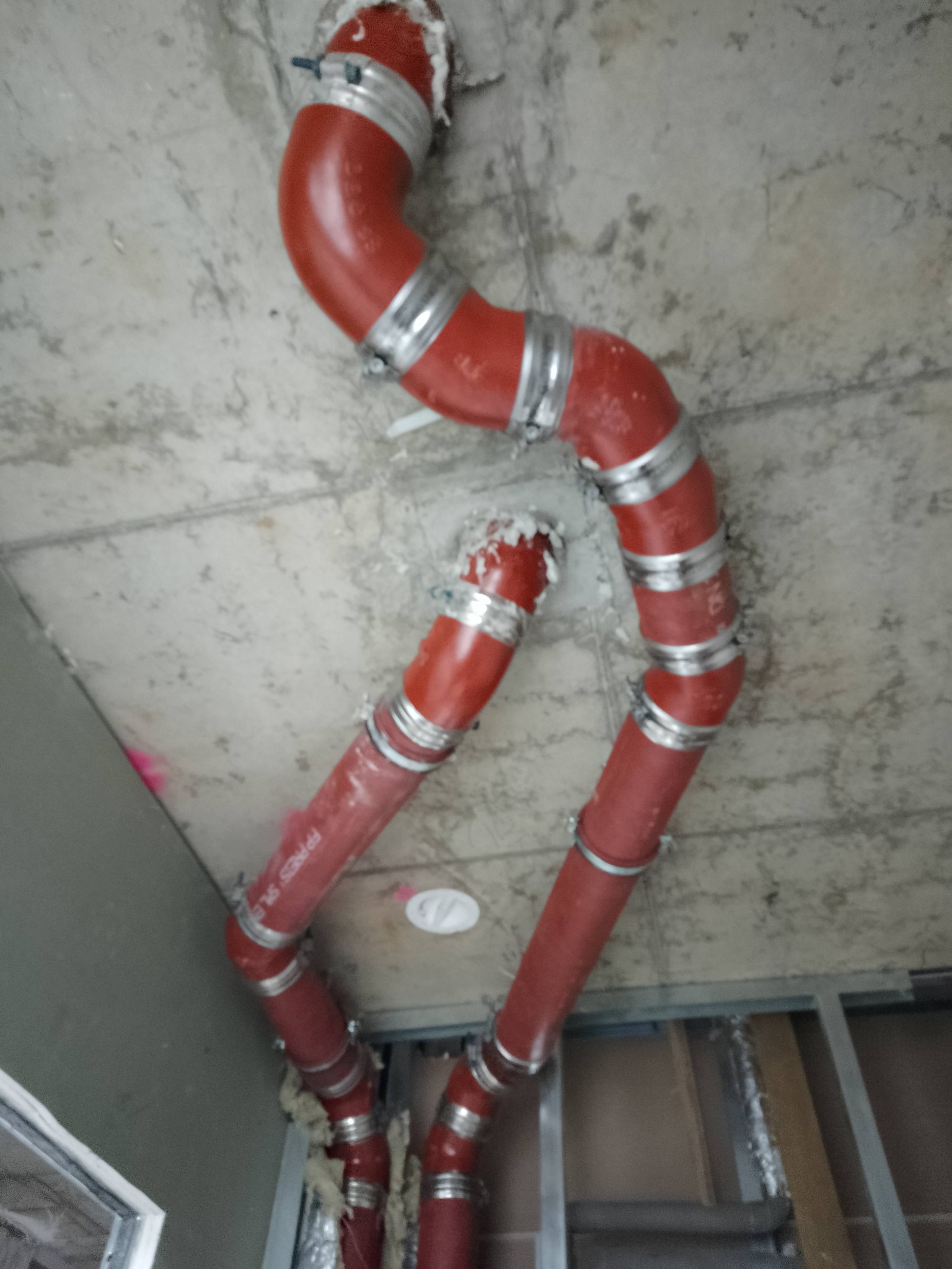 Czerwone rury instalacyjne z metalowymi obejmami, zamontowane pod surowym betonowym sufitem w trakcie prac wykończeniowych.