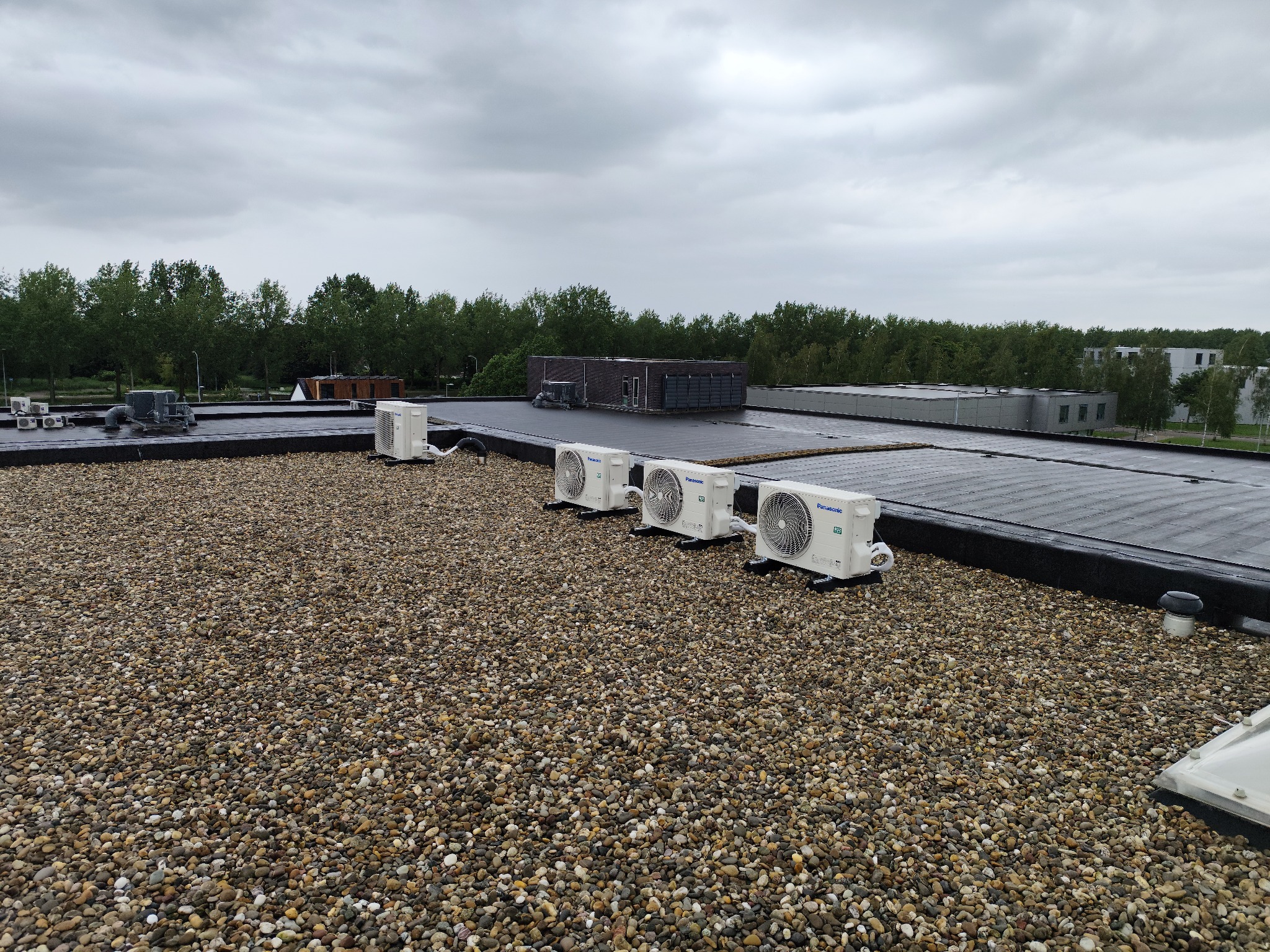 Cztery zewnętrzne jednostki klimatyzacyjne Panasonic zamontowane na płaskim dachu pokrytym żwirem, widok z góry w pochmurny dzień.