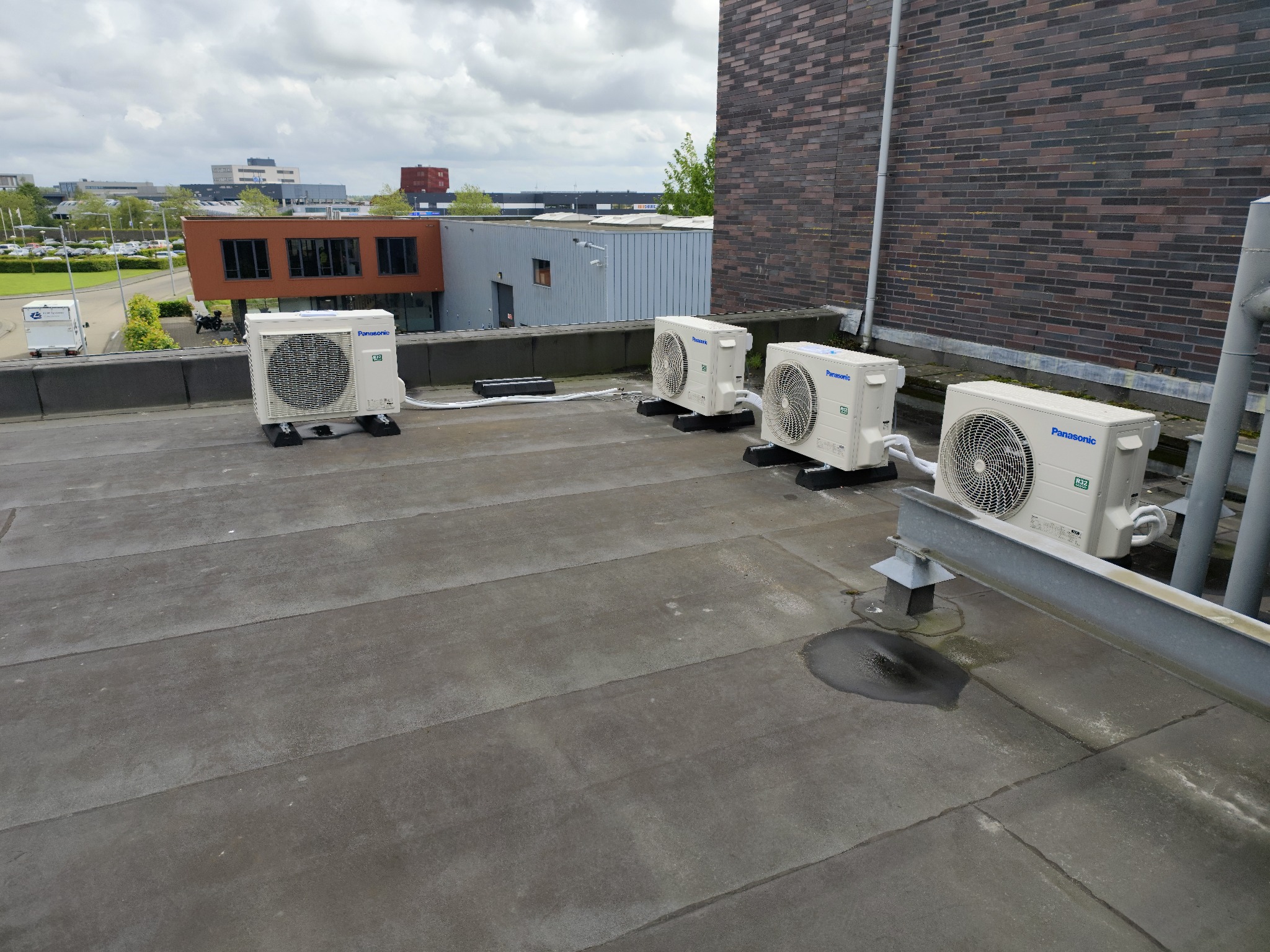 Cztery jednostki zewnętrzne klimatyzacji Panasonic zamontowane na płaskim dachu budynku z widokiem na zabudowania miejskie w pochmurny dzień.