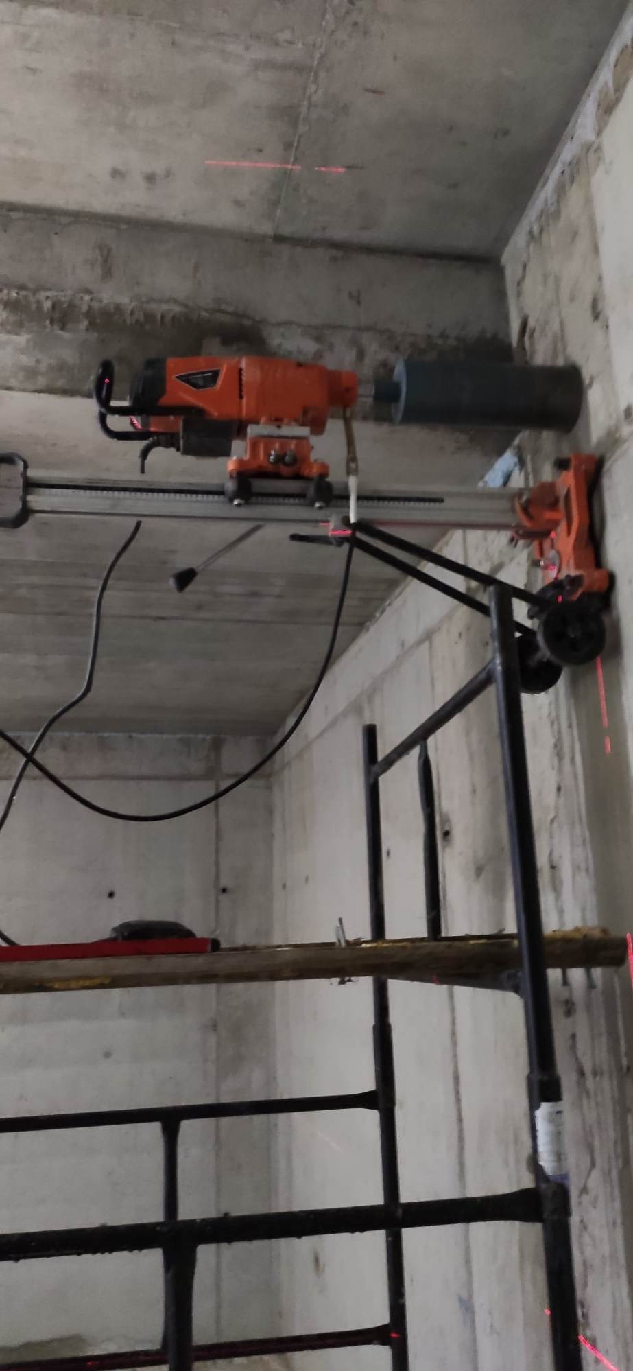 Wiertnica diamentowa, pomarańczowa, mocowana do ściany z betonu za pomocą szyny, na rusztowaniu, z widocznymi laserowymi liniami wyznaczającymi wiercenie.