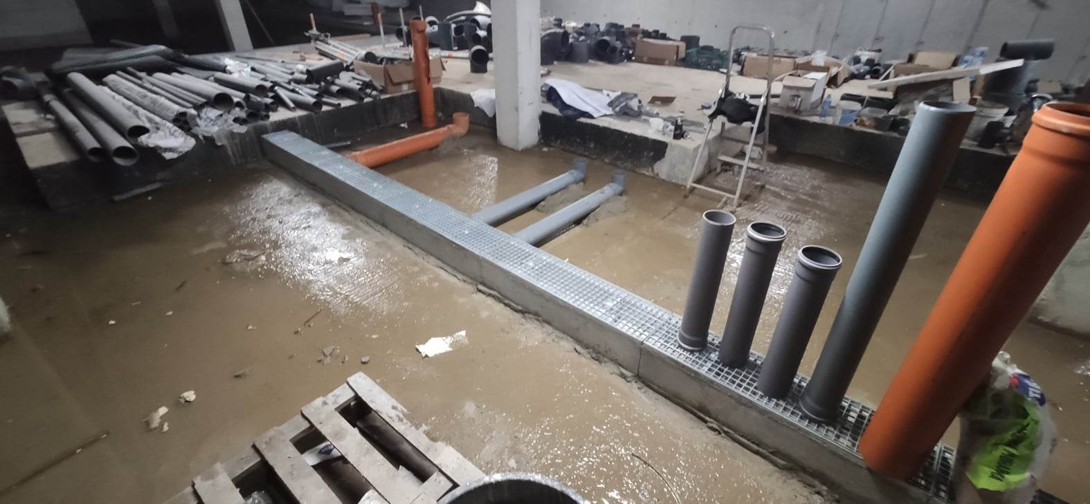 Instalacja kanalizacyjna w trakcie budowy: widoczne rury kanalizacyjne z tworzywa sztucznego w wykopie z metalową kratą, zalane wodą, w tle porozrzucane materiały budowlane i drabina.
