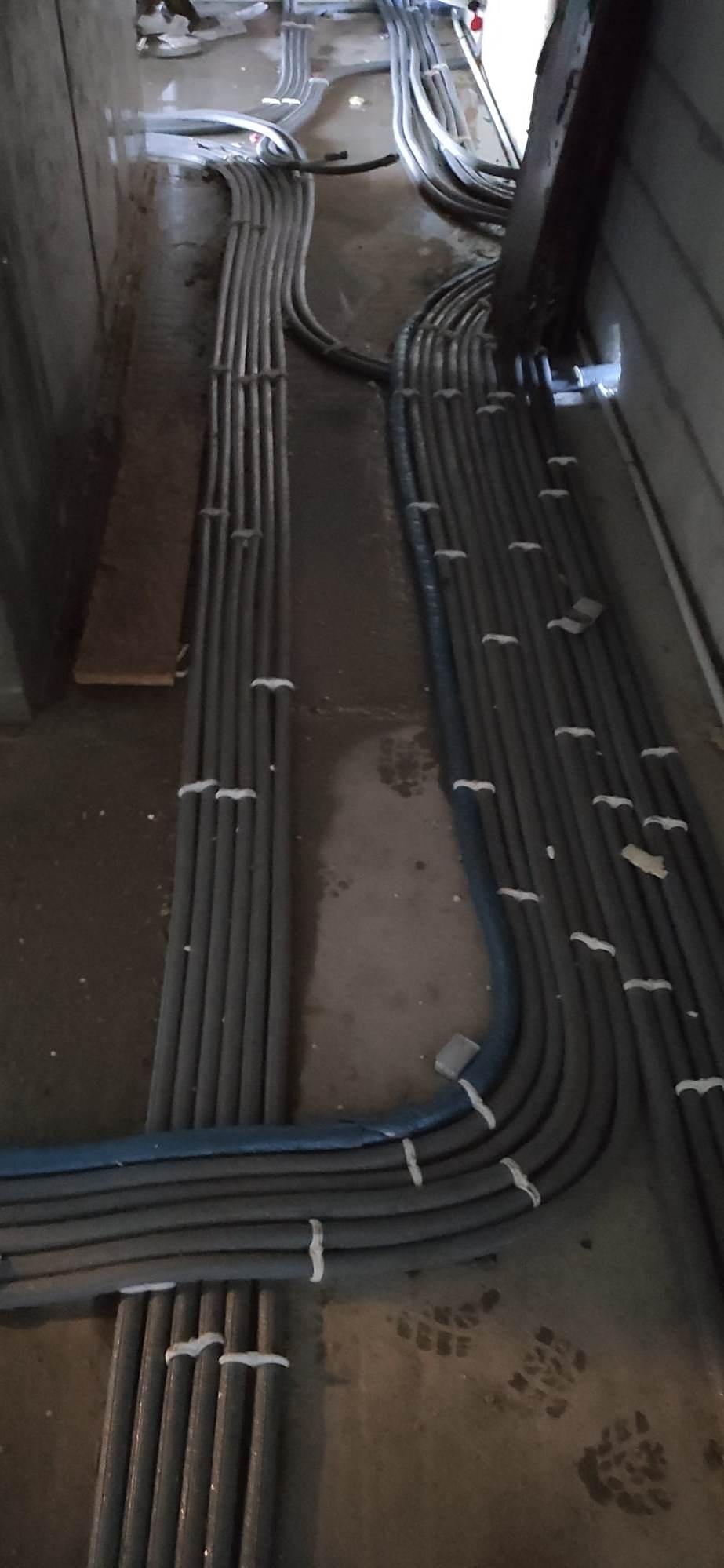 Rzędy szarych i niebieskich rur, zabezpieczonych białymi opaskami, ułożone równolegle na betonowej podłodze w surowym wnętrzu. Ślady butów widoczne na podłodze.