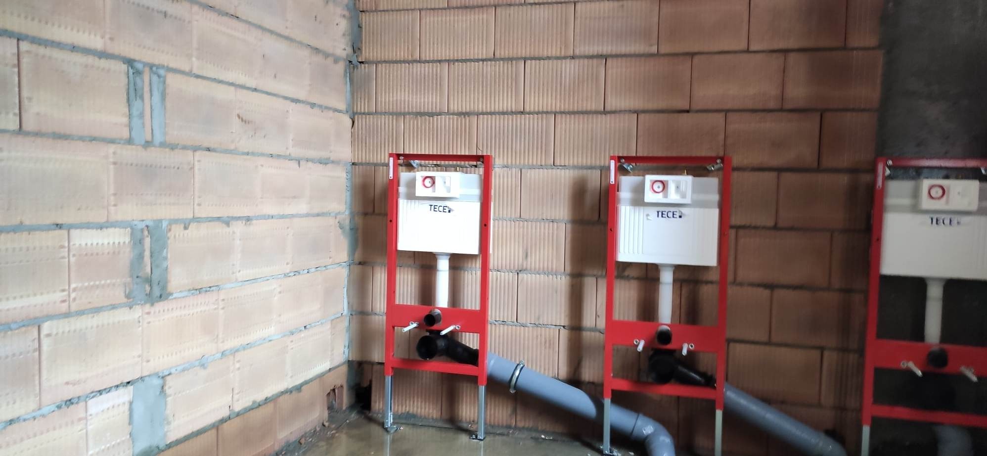 Trzy czerwone stelaże podtynkowe marki TECE zamontowane na ścianie z cegły dziurawki w surowym stanie, widoczne szare rury kanalizacyjne i czarne rury doprowadzające wodę.