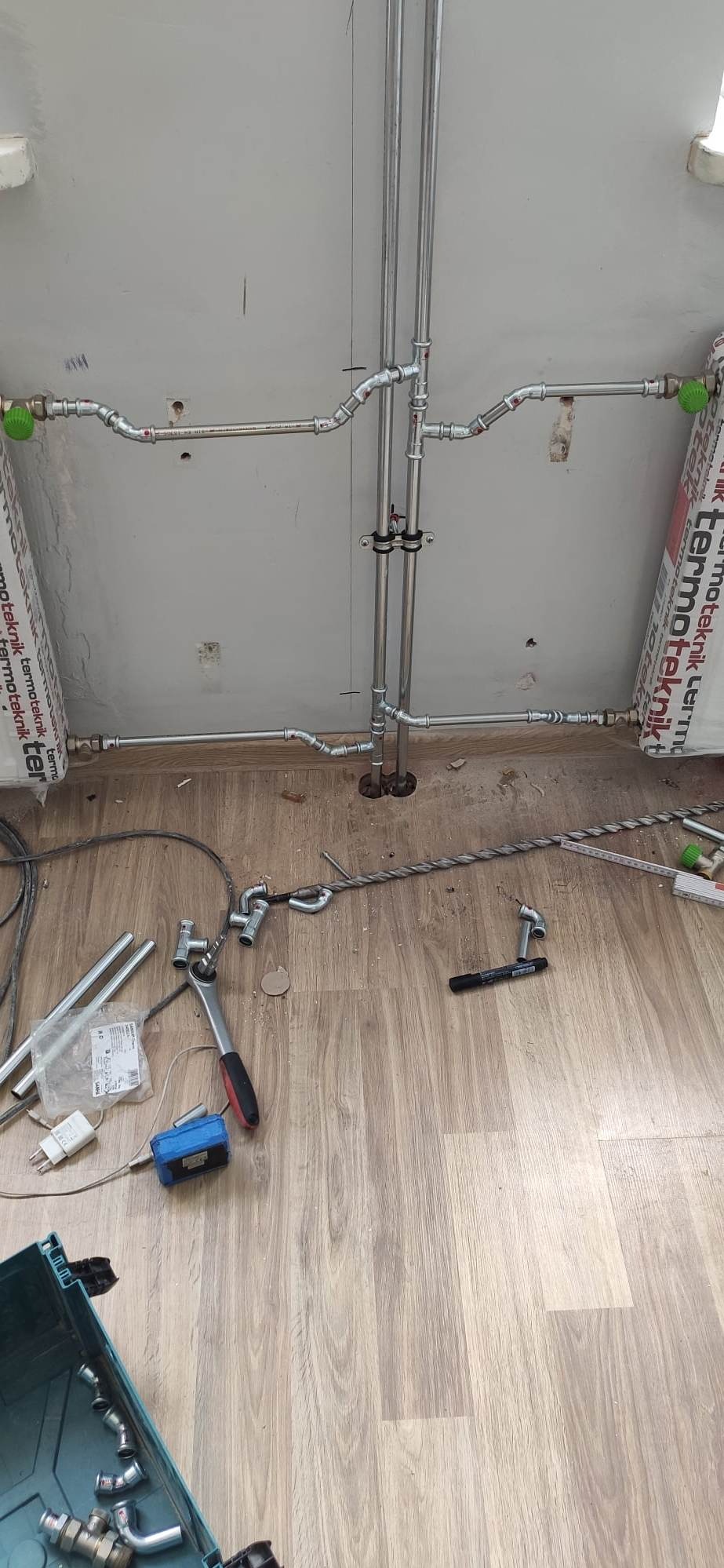 Montaż instalacji centralnego ogrzewania z rur stalowych, widoczne narzędzia i elementy instalacji na podłodze z paneli.