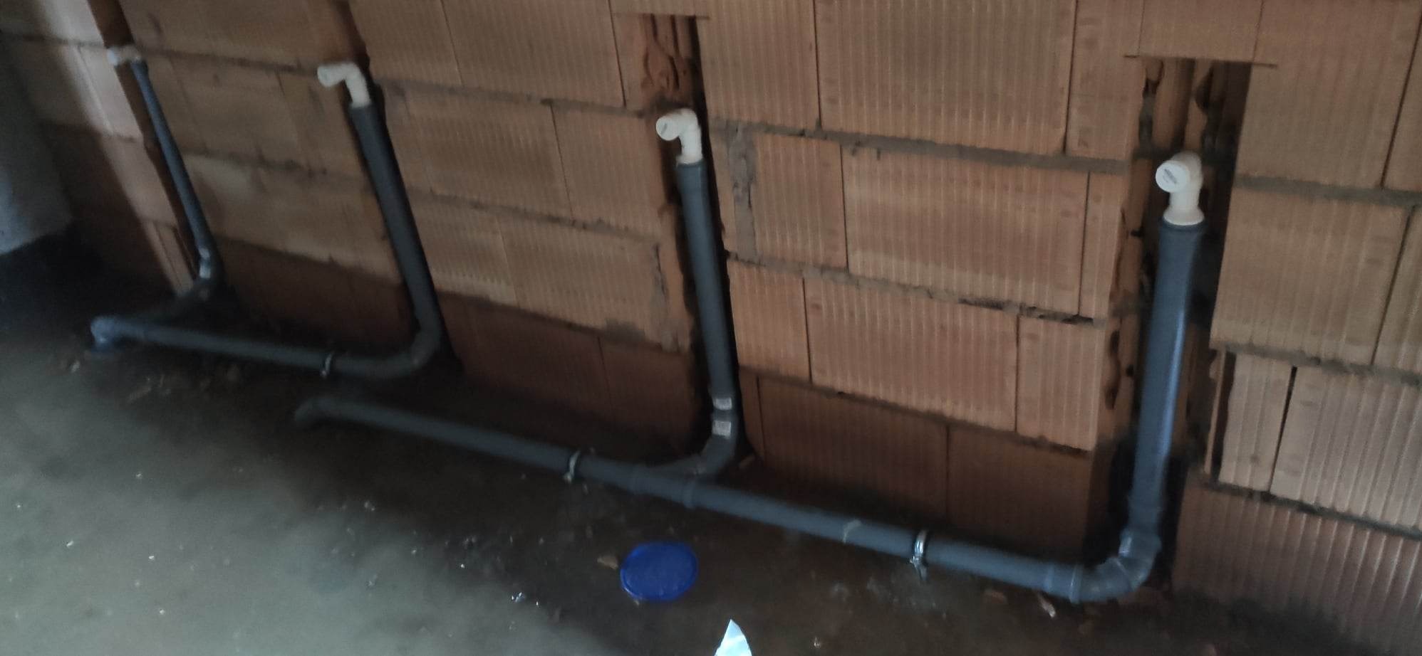 Szare rury instalacji wodnej wystające ze ściany z czerwonej cegły, widoczne łączenia i fragment niebieskiej pokrywki na podłodze.