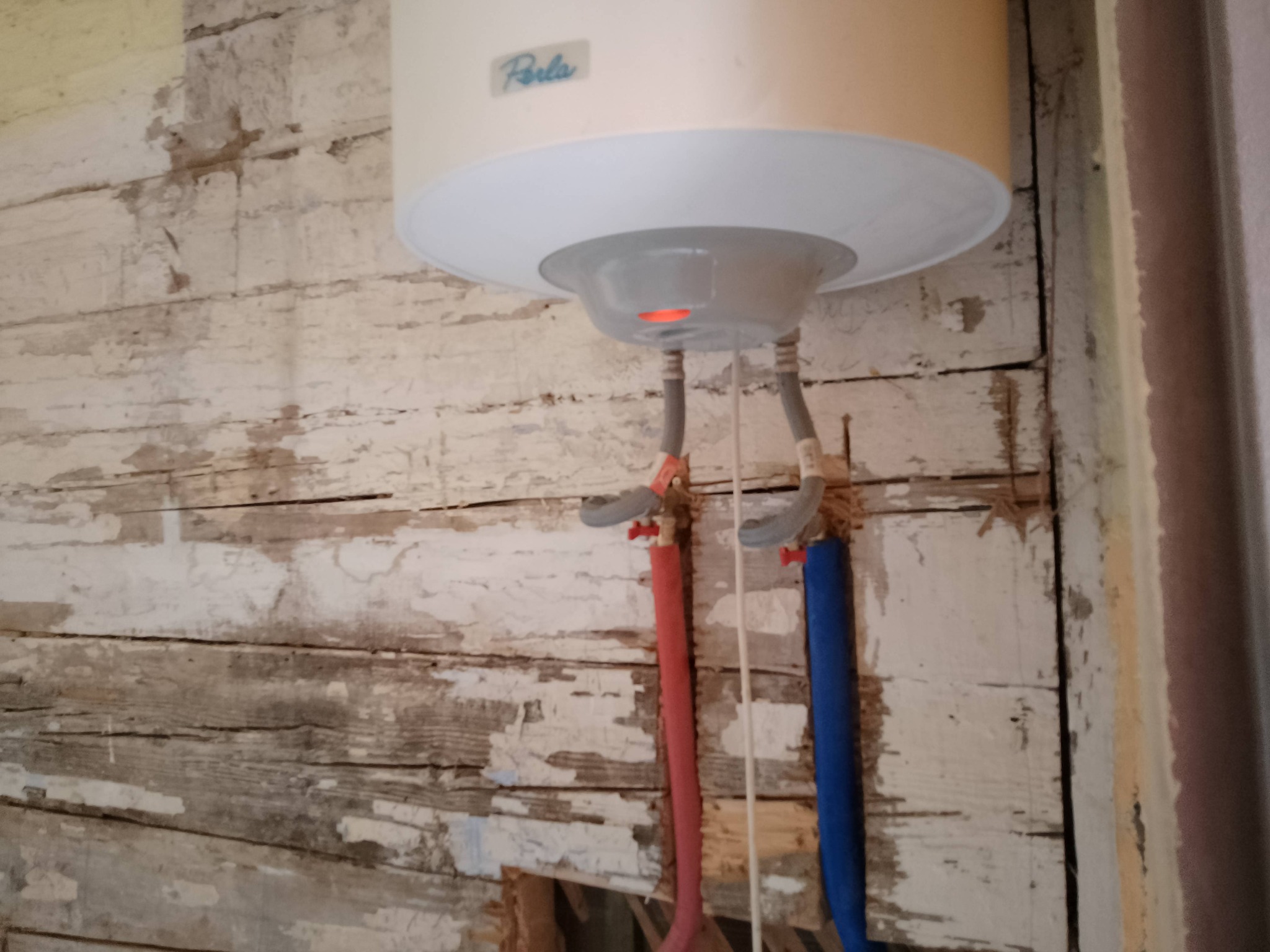 Bojler marki Perla zamontowany na starej, drewnianej ścianie z odchodzącą farbą, widoczne rury doprowadzające wodę w czerwonej i niebieskiej izolacji.