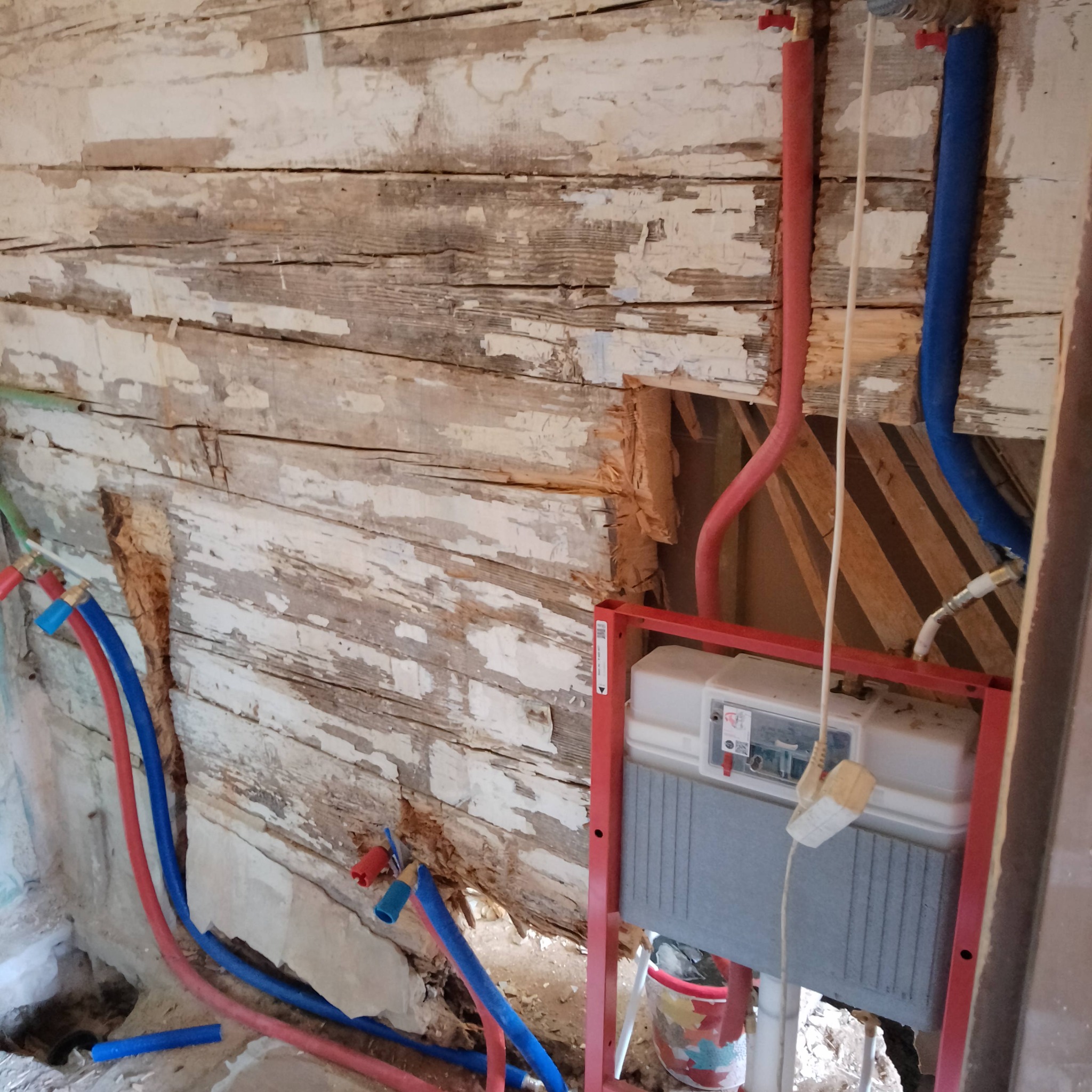 Stelaż podtynkowy WC zamontowany na tle starej, drewnianej ściany z odpadającą farbą, widoczne rury doprowadzające wodę w kolorach czerwonym i niebieskim.