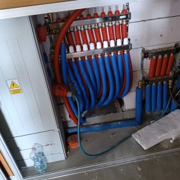 Radosław Saberniak INSTAL-RAD - Rozdzielacz instalacji grzewczej z czerwono-niebieskimi rurami, zamontowany na ścianie z bloczków, widoczna skrzynka z ostrzeżeniem elektrycznym i butelka wody na podłodze.