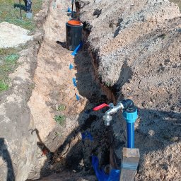 Radosław Saberniak INSTAL-RAD - Wykop z widocznymi fragmentami instalacji wodnej, w tym rura z zaworem tymczasowo podparta drewnianym klockiem, oraz czarny zbiornik w ziemi, prawdopodobnie na szambo lub oczyszczalnię, teren...