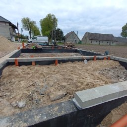 Radosław Saberniak INSTAL-RAD - Wykop pod fundamenty domu jednorodzinnego z izolacją przeciwwilgociową, częściowo zasypany piaskiem, z widocznymi rurami kanalizacyjnymi i szpadlem. W tle zabudowa wiejska i samochód dostawczy.