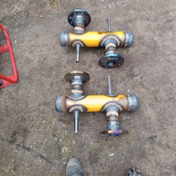 Radosław Saberniak INSTAL-RAD - Dwa żółte, spawane kolektory rurowe z kołnierzami, leżące na ziemi obok butów roboczych, widoczne ślady spawania i przypalenia farby.