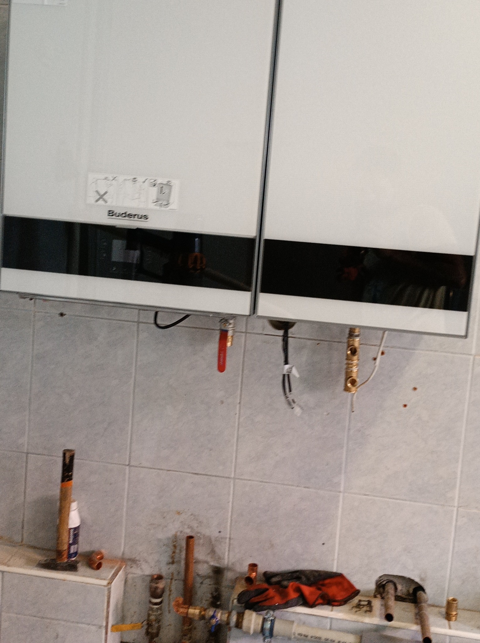Dwa wiszące, białe piece gazowe marki Buderus, z widocznymi przyłączami hydraulicznymi i gazowymi w trakcie montażu, oraz narzędzia i akcesoria hydrauliczne na tle kafelkowanej ściany.