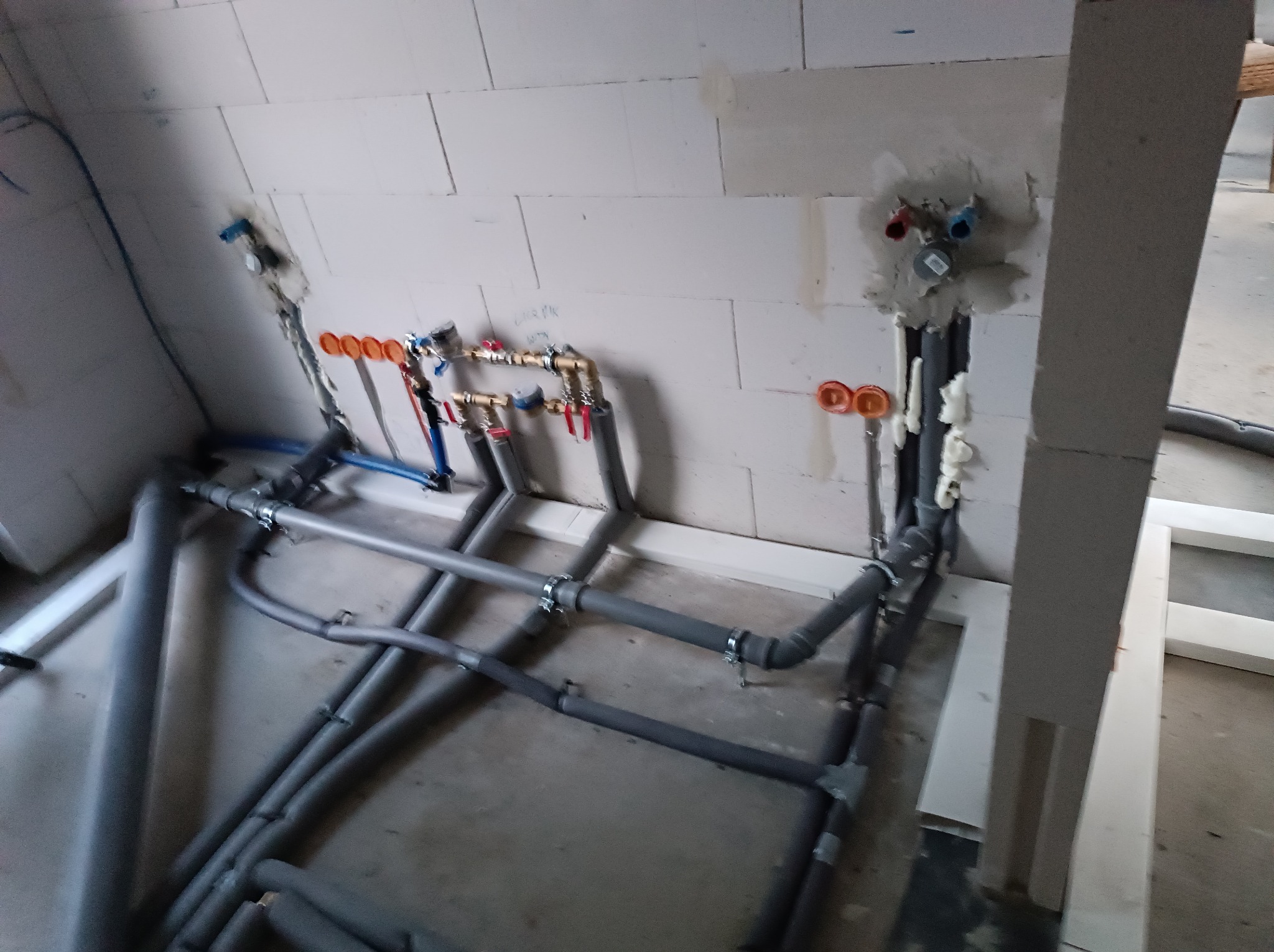 Szare rury instalacji wodnej i grzewczej w surowym wnętrzu, widoczne przyłącza wody ciepłej i zimnej w ścianie z betonu komórkowego, częściowo ocieplone.