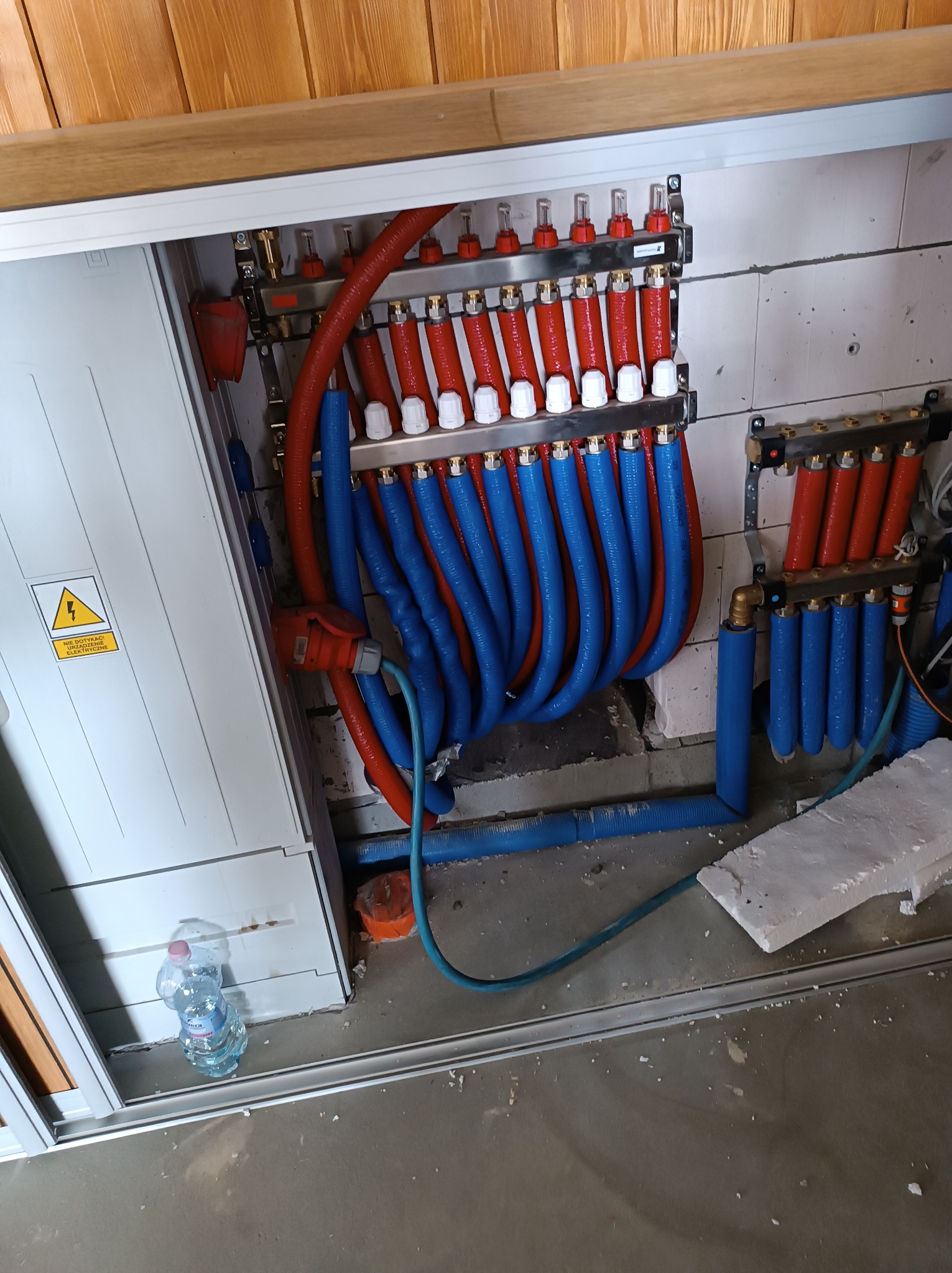 Rozdzielacz instalacji grzewczej z czerwono-niebieskimi rurami, zamontowany na ścianie z bloczków, widoczna skrzynka z ostrzeżeniem elektrycznym i butelka wody na podłodze.