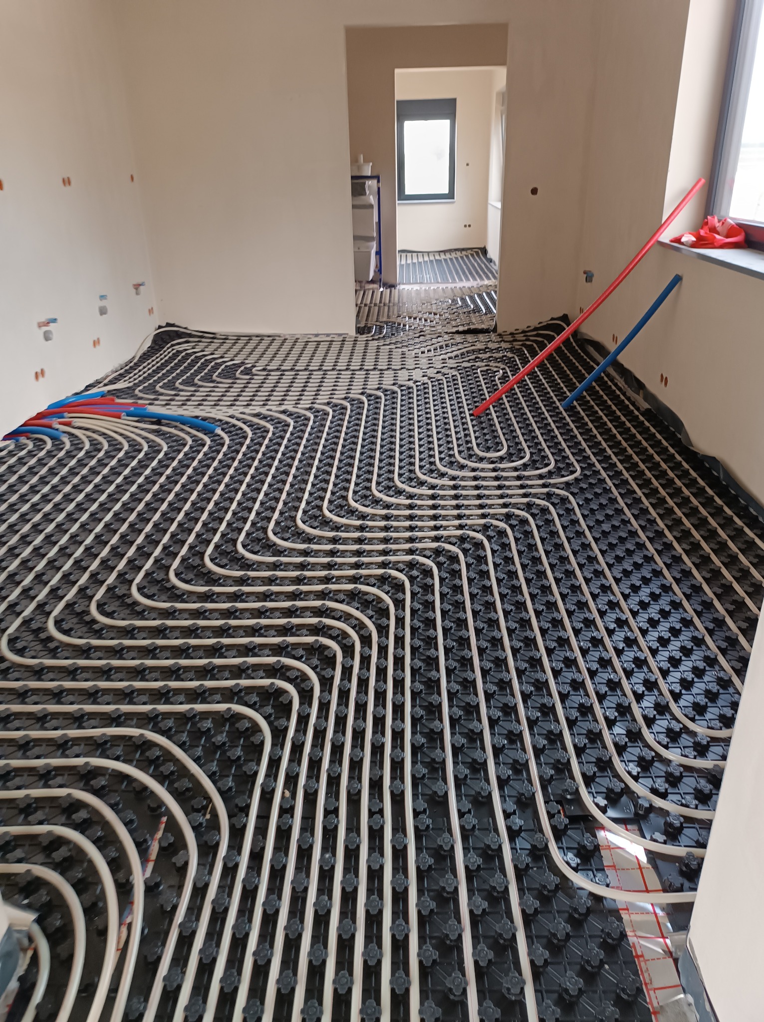 Rozłożona instalacja ogrzewania podłogowego z rurkami w kształcie meandrów na czarnej macie izolacyjnej, widoczne czerwone i niebieskie rurki doprowadzające w tle.