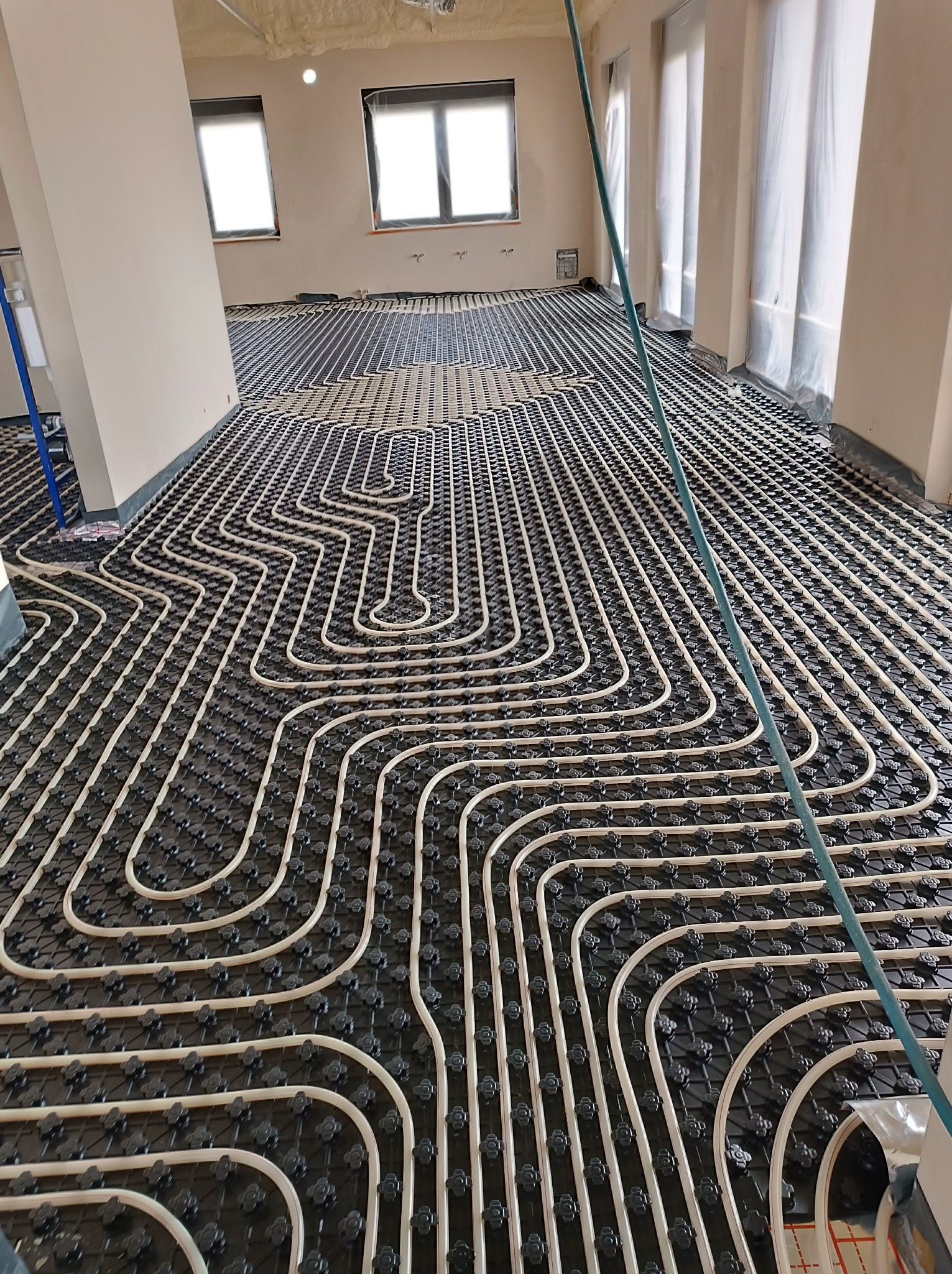 Rozłożona instalacja ogrzewania podłogowego z białych rurek na czarnej macie w surowym wnętrzu, z oknami zabezpieczonymi folią malarską.