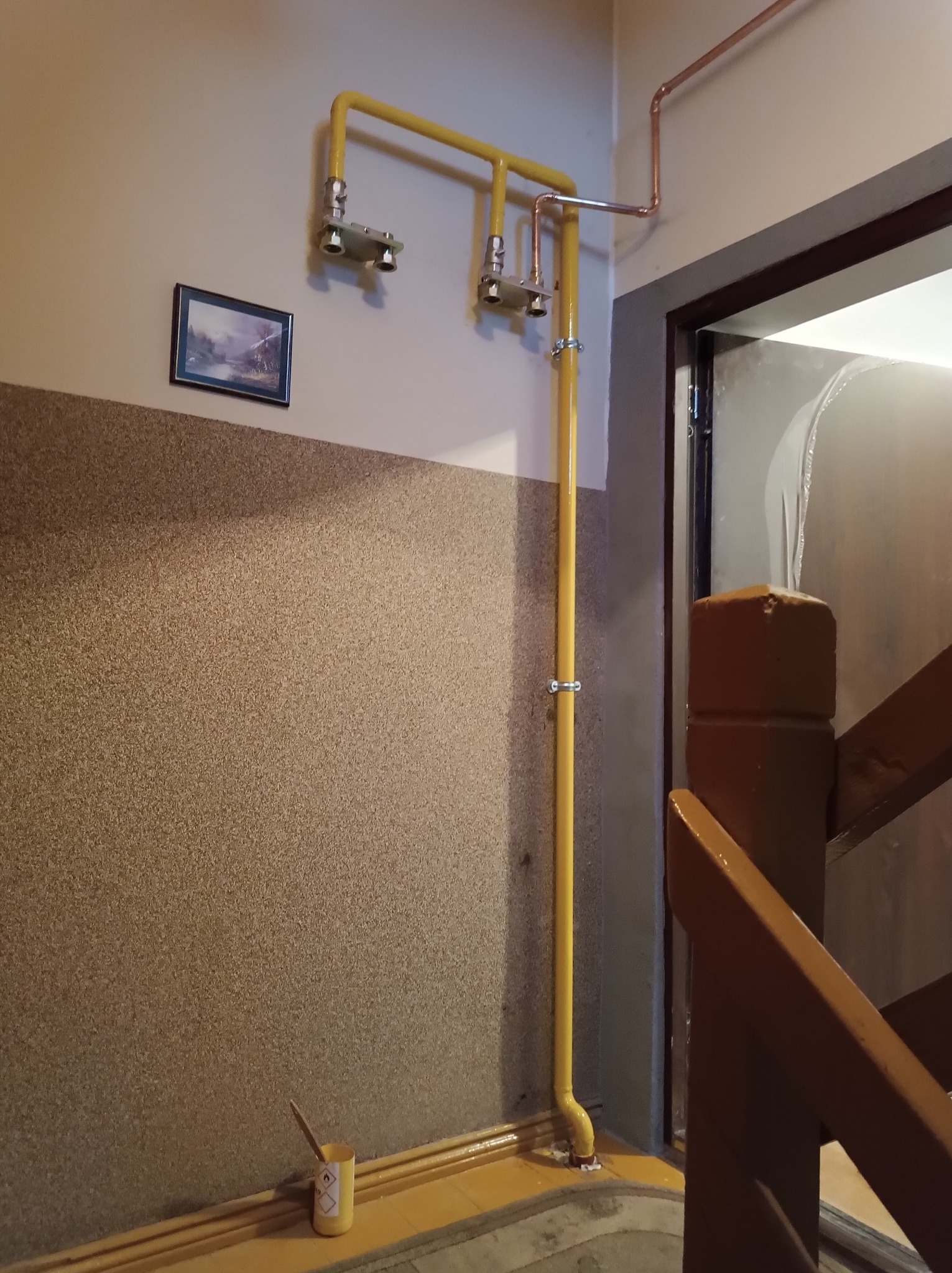 Żółta instalacja gazowa z dwoma zaworami, połączona z miedzianą instalacją, zamontowana na ścianie w korytarzu. Na ścianie wisi obrazek, a na podłodze stoi puszka z farbą.