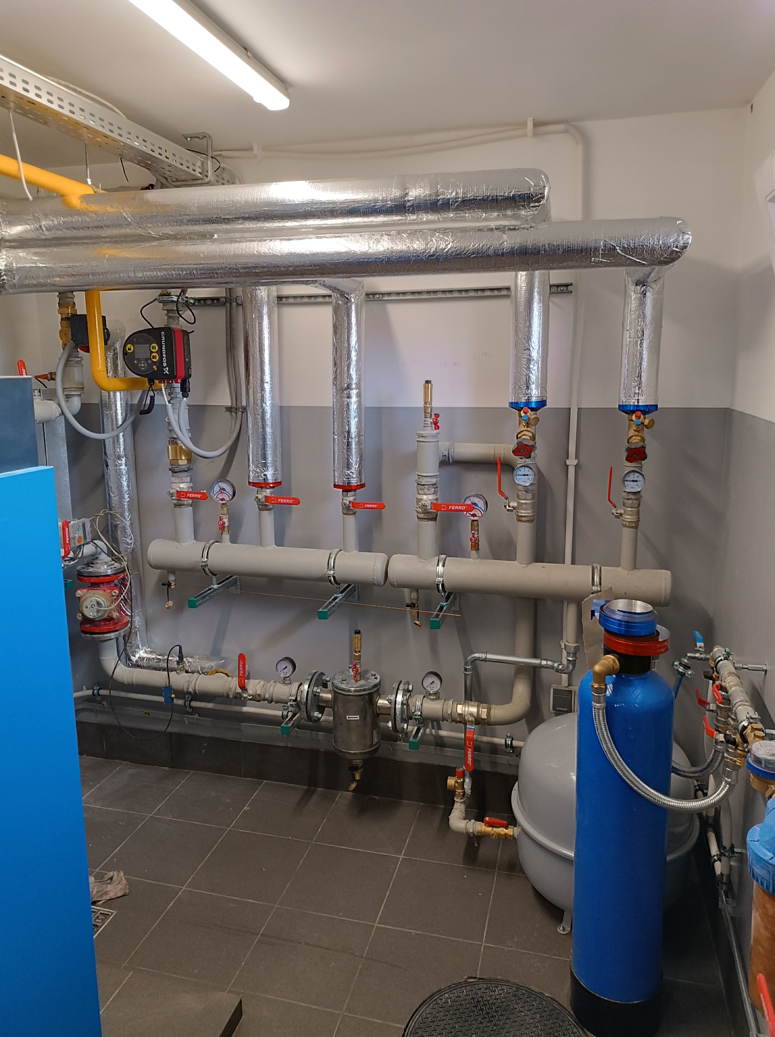 Szczegółowe ujęcie instalacji grzewczej z izolowanymi rurami, zaworami Ferro, pompą obiegową Grundfos, manometrami i filtrami, w pomieszczeniu technicznym z szarą podłogą.