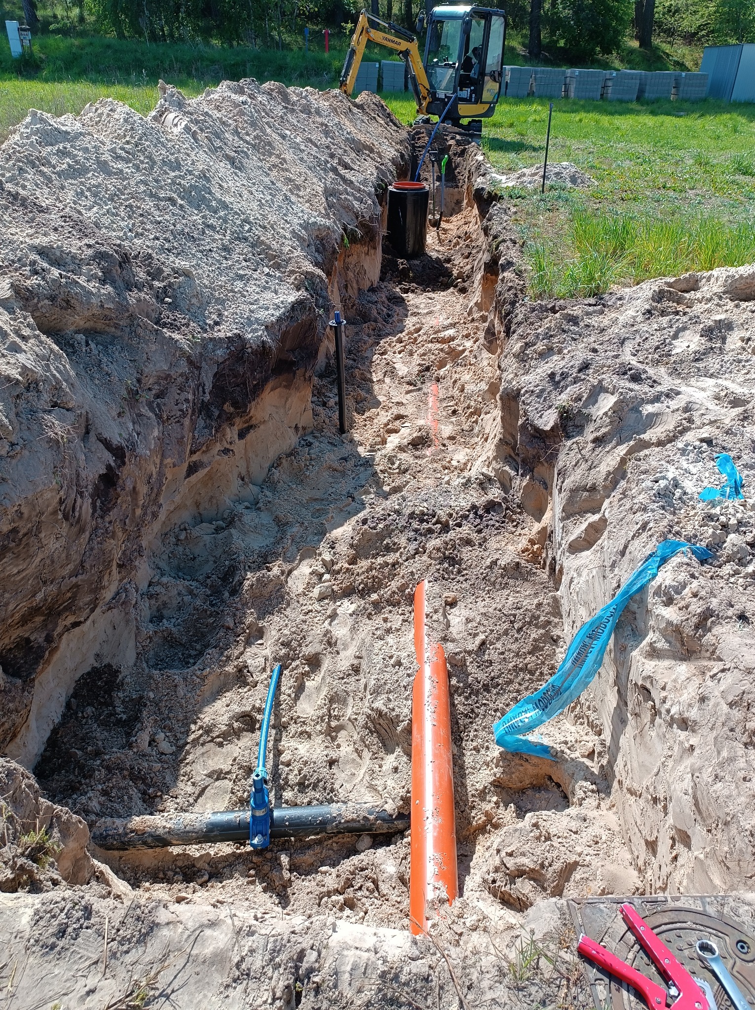 Wykop w ziemi z widocznymi rurami: pomarańczową kanalizacyjną i czarną z zaworem, niebieska taśma ostrzegawcza, mini koparka w tle.