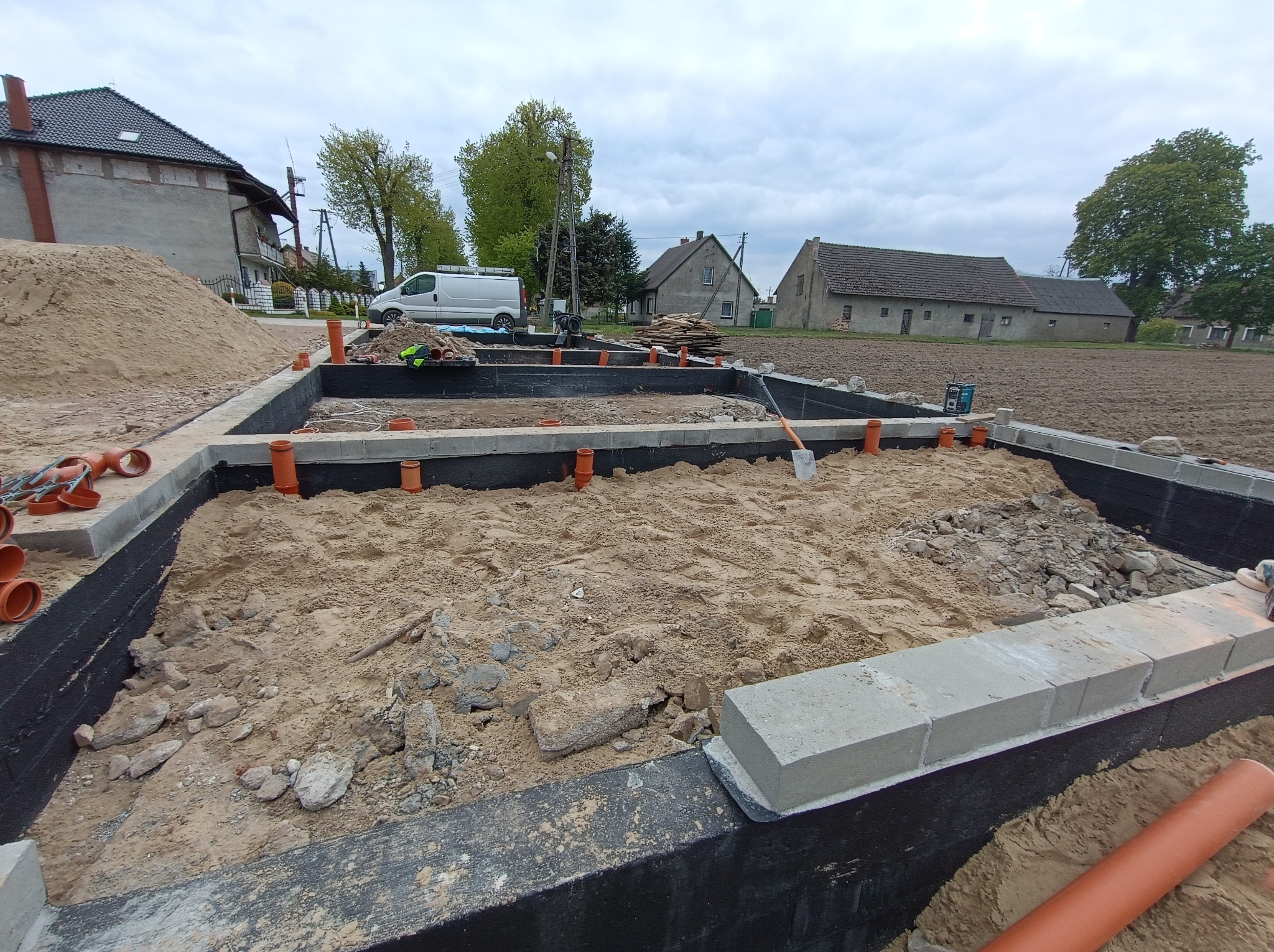 Wykop pod fundamenty domu jednorodzinnego z izolacją przeciwwilgociową, częściowo zasypany piaskiem, z widocznymi rurami kanalizacyjnymi i szpadlem. W tle zabudowa wiejska i samochód dostawczy.