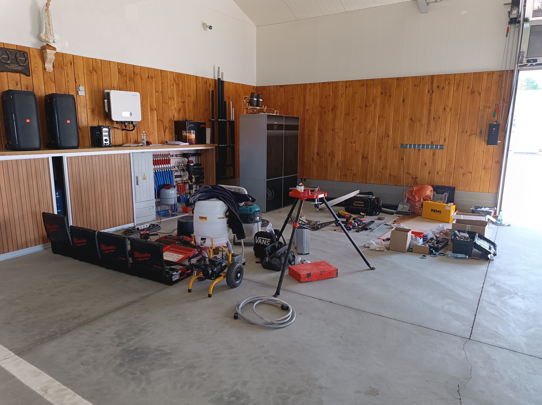 Rozległe pomieszczenie z drewnianą boazerią, zawierające piec grzewczy, rozdzielnicę hydrauliczną, narzędzia Milwaukee, agregat malarski, skrzynkę narzędziową i akcesoria hydrauliczne na betonowej...