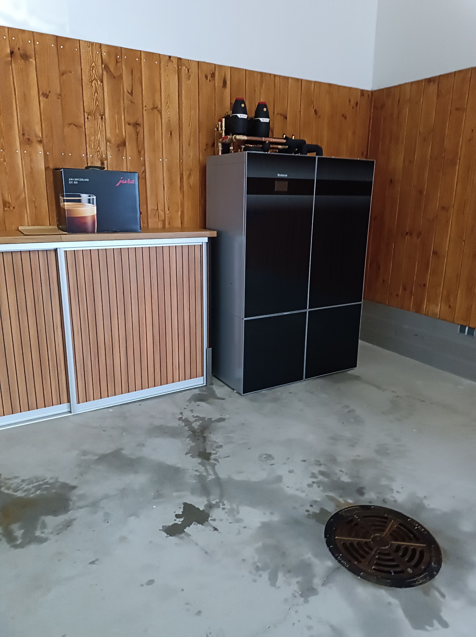 Nowoczesny, dwuczęściowy piec gazowy Buderus z miedzianymi rurami, zainstalowany w pomieszczeniu z drewnianą boazerią i szafką. Na szafce stoi pudełko po kawie Jura. Na podłodze widoczny właz...