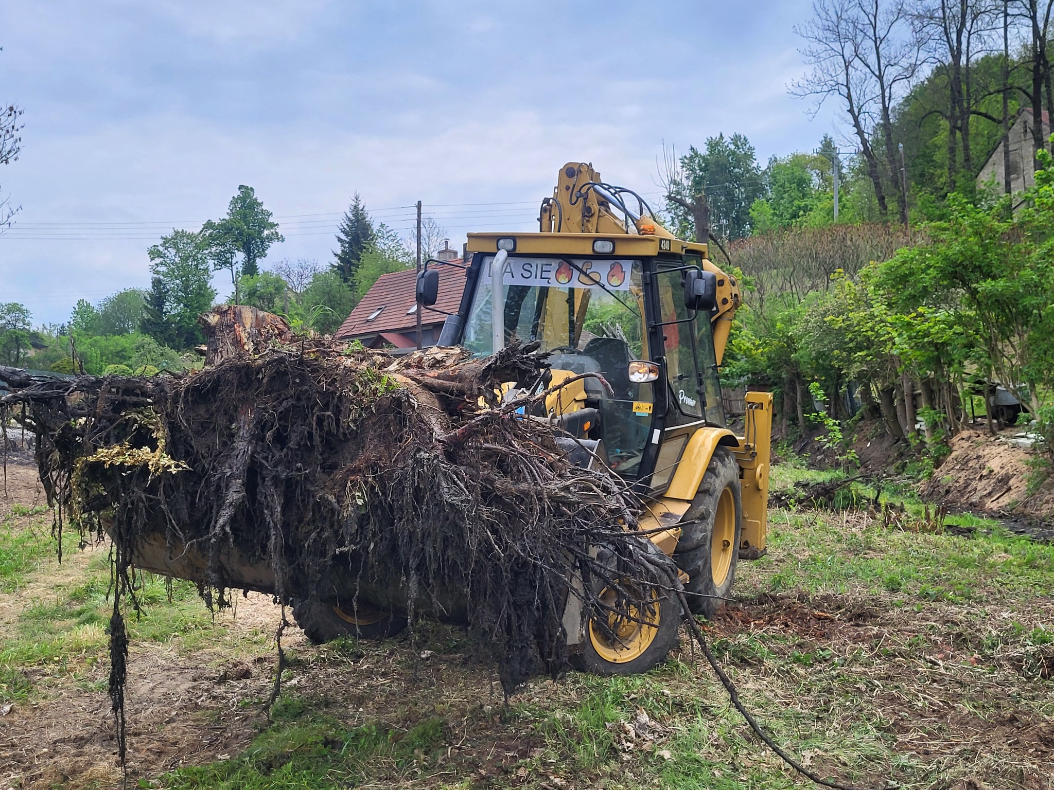 Żółta koparko-ładowarka z pełną łyżką wykopanych korzeni drzew, na przedniej szybie napis 'DA SIĘ' z emotikonami ognia, w tle dom z czerwonym dachem i drzewa.