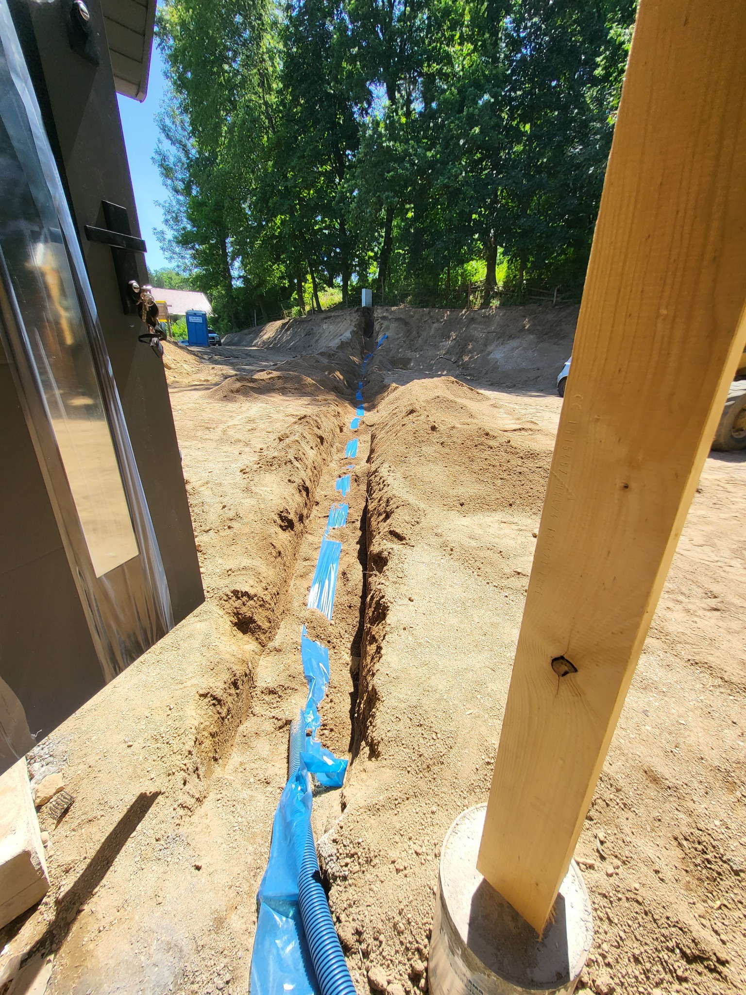 Długi wykop w piaszczystej ziemi z ułożoną niebieską rurą karbowaną w niebieskiej folii sygnalizacyjnej, widok z perspektywy ziemi, w tle drzewa i niebieska toaleta przenośna.
