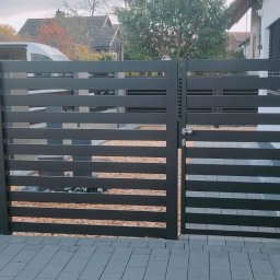 STEEL WINGS MARKUS REISER - Nowoczesna brama wjazdowa z poziomych, czarnych profili, z dekoracyjną dynią Halloween obok słupka, na tle brukowanej posesji i zabudowy jednorodzinnej.