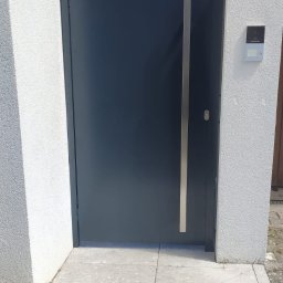 STEEL WINGS MARKUS REISER - Nowoczesna, grafitowa brama wjazdowa z długim, pionowym uchwytem ze stali nierdzewnej, osadzona między białymi, teksturowanymi słupkami, z widocznym domofonem i fragmentem chodnika z kostki.