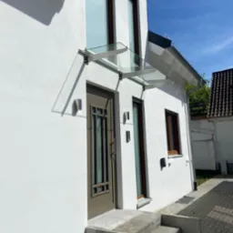 Wejście do nowoczesnego domu z szarobrązowymi drzwiami i szklanym zadaszeniem wspartym na metalowych wspornikach, oświetlone bocznym światłem słonecznym, z widocznymi schodami wejściowymi...