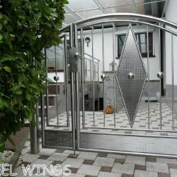 STEEL WINGS MARKUS REISER - Nowoczesna, dwuskrzydłowa brama ze stali nierdzewnej z dekoracyjnym rombem i kulistymi elementami, widok z zewnątrz, z boku zielony krzew.