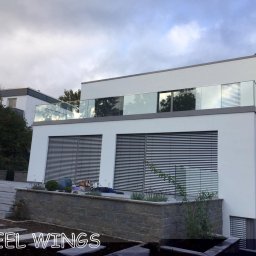 STEEL WINGS MARKUS REISER - Nowoczesny dom z balkonem zabezpieczonym szklaną konstrukcją, widoczne żaluzje w oknach i fragment ogrodu z kamiennym murkiem.