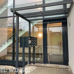 STEEL WINGS MARKUS REISER - Nowoczesne wejście do budynku z przeszklonymi drzwiami i zadaszeniem, skrzynki pocztowe na metalowym stelażu, widok na schody wewnętrzne przez duże okno.