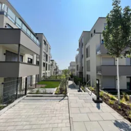 Nowoczesne osiedle z balkonami z przeszklonymi barierkami, niskimi metalowymi ogrodzeniami i brukowanym chodnikiem w słoneczny dzień.