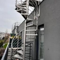 Zewnętrzne schody spiralne ze stali nierdzewnej z balustradą, prowadzące na dach budynku z szarą elewacją i widocznym oknem.