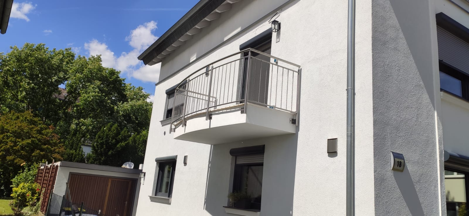 Nowoczesny budynek z białym tynkiem i balkonem z zaokrągloną, metalową balustradą, widoczne rolety okienne i numer domu 18.