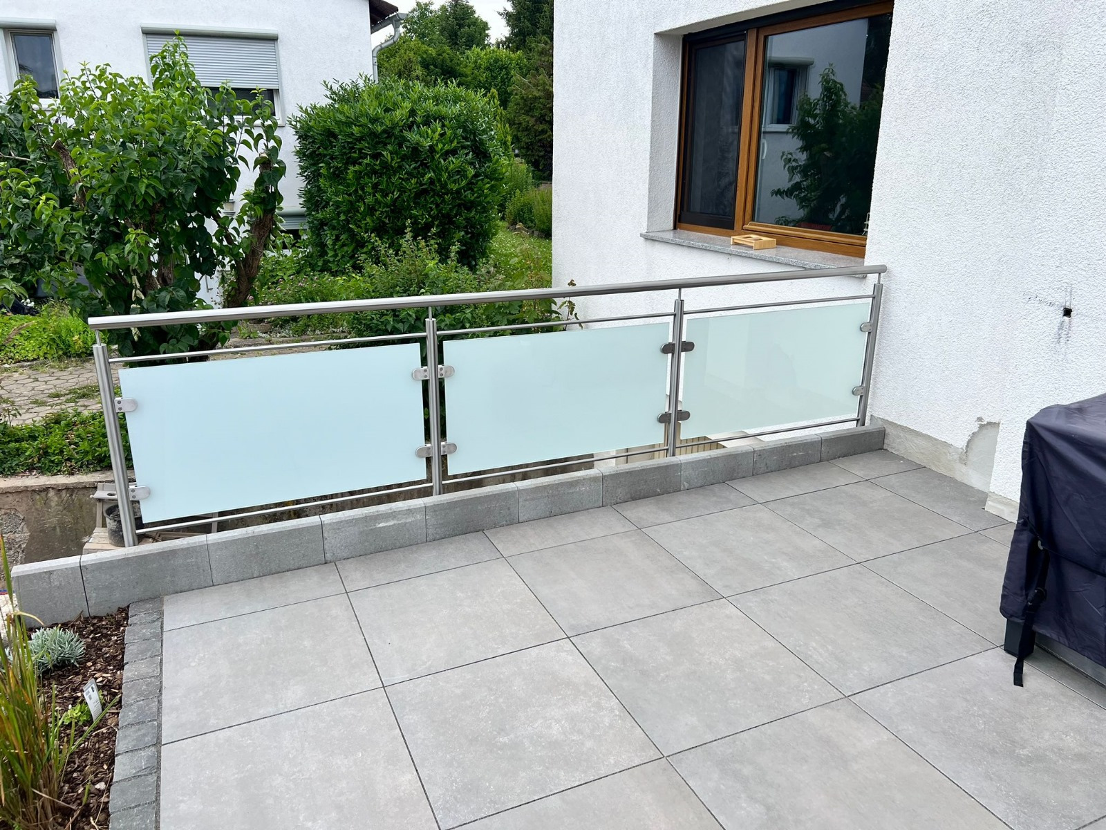 Nowoczesna balustrada ze stali nierdzewnej z mlecznymi panelami szklanymi, zamontowana na tarasie wyłożonym szarą kostką brukową, z widokiem na zielony ogród.