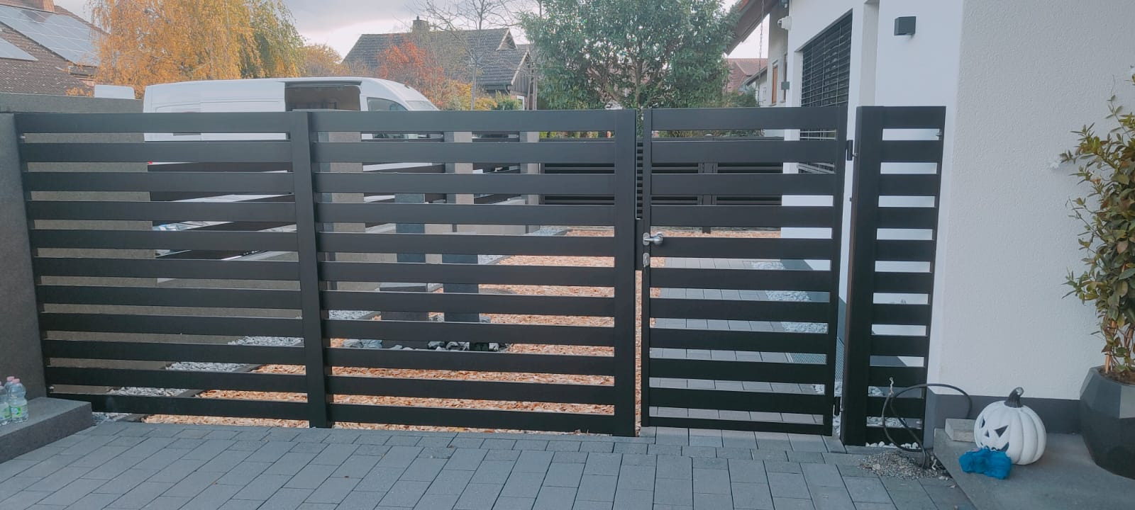 Nowoczesna brama wjazdowa z poziomych, czarnych profili, z dekoracyjną dynią Halloween obok słupka, na tle brukowanej posesji i zabudowy jednorodzinnej.