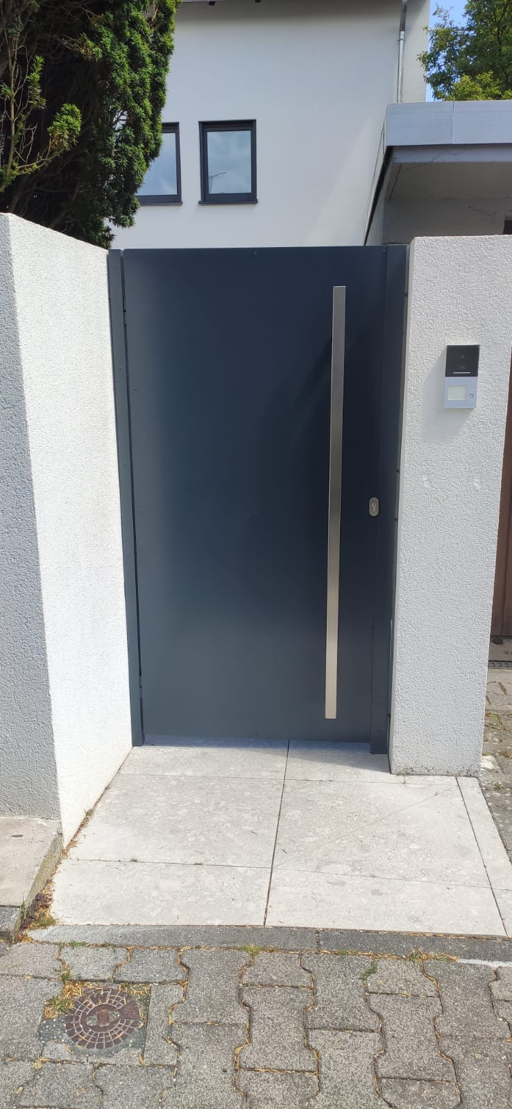 Nowoczesna, grafitowa brama wjazdowa z długim, pionowym uchwytem ze stali nierdzewnej, osadzona między białymi, teksturowanymi słupkami, z widocznym domofonem i fragmentem chodnika z kostki.